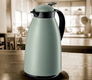 Grüne matte Isolierkanne mit schwarzem Griff und Deckel, auf einem Tisch in einem Café aus seitlicher Perspektive.