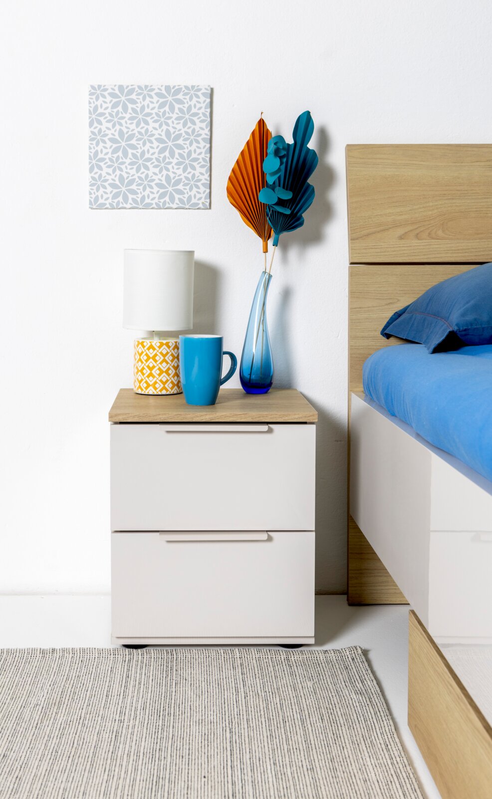 Moderner weißer Nachttisch mit zwei Schubladen und Holzplatte neben einem Bett, dekoriert mit Tischlampe, blauer Tasse und blauer Glasvase mit orange-blauen Trockenblättern; frontale Ansicht auf Augenhöhe.