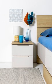 Moderne witte nachtkastje met twee lades en een houten blad naast een bed, versierd met een tafellamp, een blauwe kop en een blauwe glazen vaas met oranje-blauwe gedroogde bladeren; vooraanzicht op ooghoogte.
