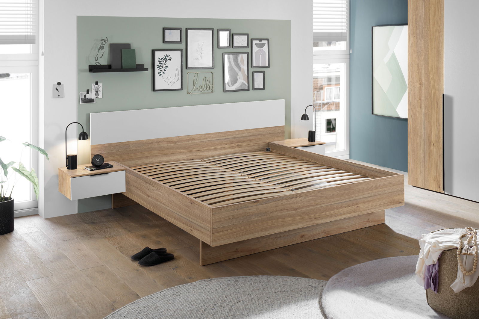 Modernes Schlafzimmer mit einem Holz-Doppelbett, das von der Seite betrachtet wird. Das Bett hat ein weißes Kopfteil und integrierte Nachttische mit Lampen. An der Wand hängen Bilderrahmen und Regale. Der Raum ist hell und stilvoll eingerichtet.