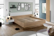 Een moderne slaapkamer met een houten tweepersoonsbed, gezien vanaf de zijkant. Het bed heeft een wit hoofdeinde en ingebouwde nachtkastjes met lampjes. Aan de muur hangen fotolijsten en planken. De kamer is licht en stijlvol ingericht.