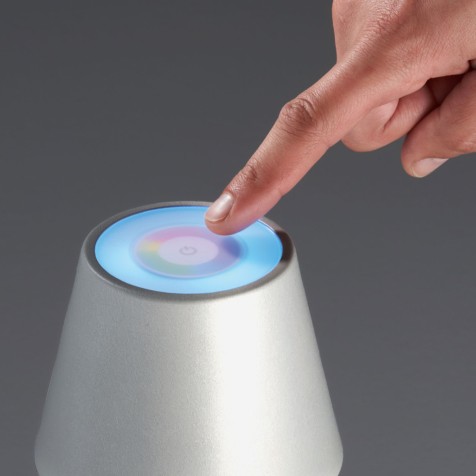 Nahaufnahme einer silbernen Akku-Tischleuchte COSENZA mit einem Finger, der den Touchscreen auf der Oberseite berührt. Der Bildschirm zeigt ein farbiges Licht und ein Power-Symbol.