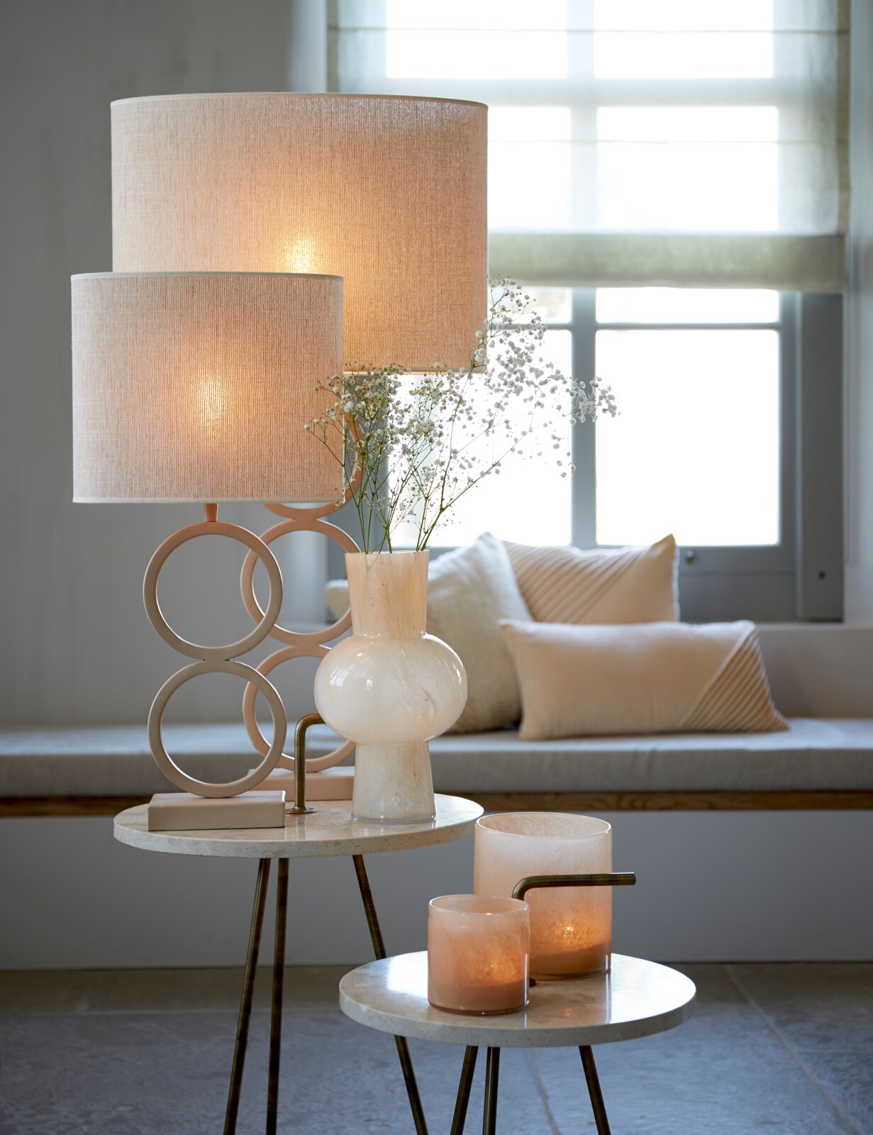 Stilvolle Tischlampe mit beigem Stoffschirm und ringförmigem Fuß auf rundem Beistelltisch, daneben weiße Glasvase mit zarten Blumen und Kerzenhalter im Wohnzimmer; Perspektive: frontale, leicht seitliche Ansicht auf Augenhöhe.
