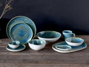 Schräg von vorn fotografiertes Keramikgeschirr-Set in Blau-Grün mit reaktiver Glasur auf Holztisch vor dunklem Hintergrund: Teller, Schalen, Platte und Tasse.