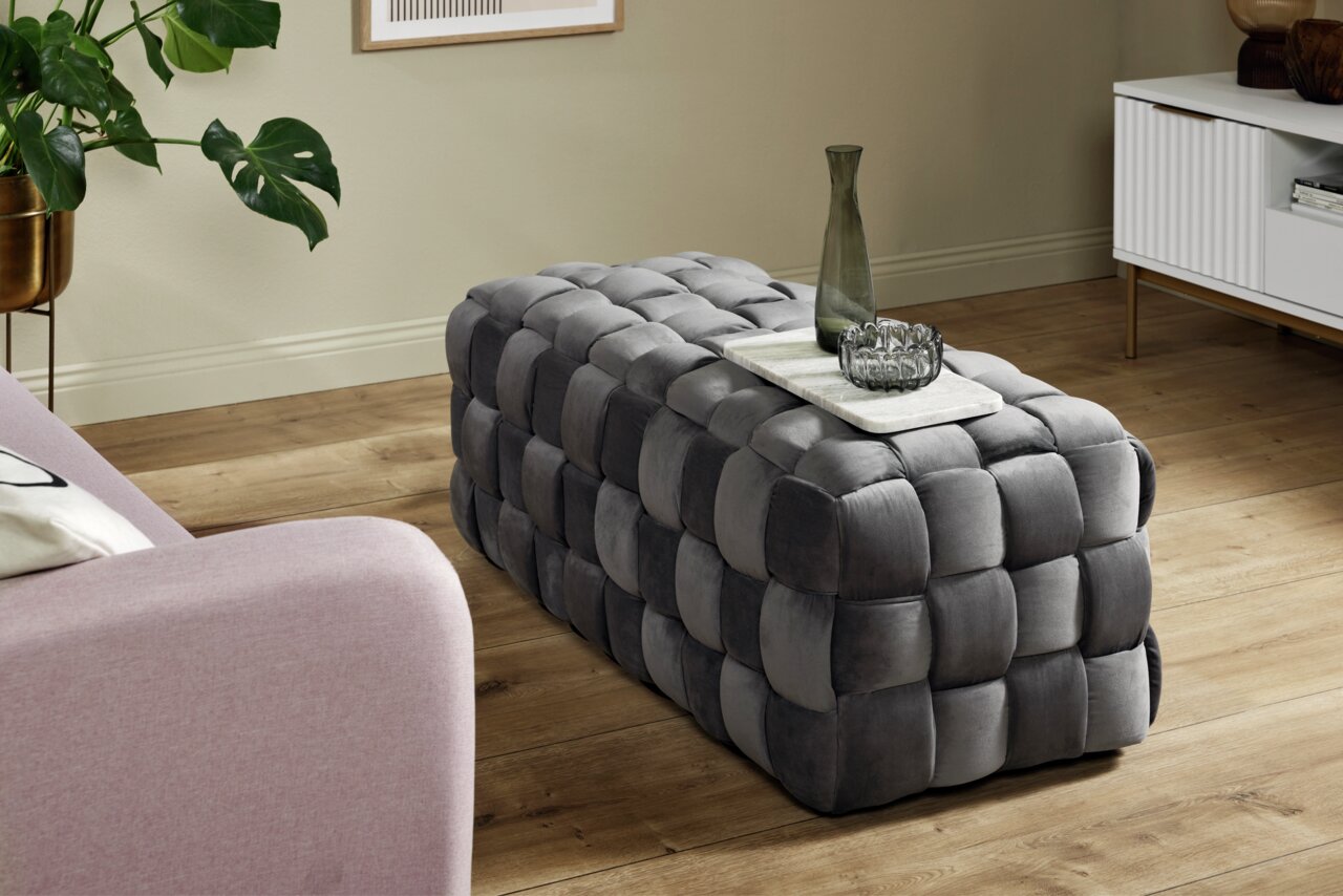 Grauer, gepolsterter Hocker in rechteckiger Form mit geflochtenem Design, seitliche Perspektive.