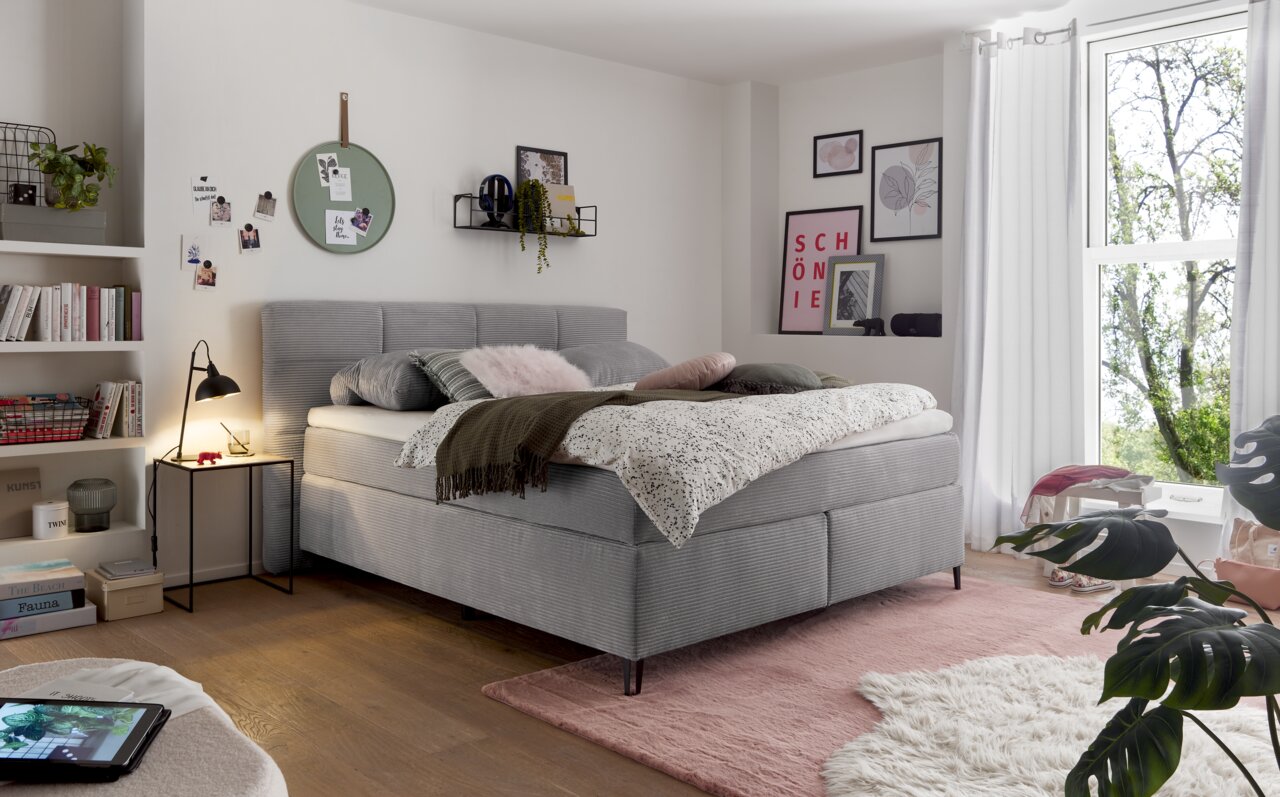 Gemütliches Boxspringbett in einem stilvoll eingerichteten Schlafzimmer, seitliche Perspektive mit Dekokissen und Decken.
