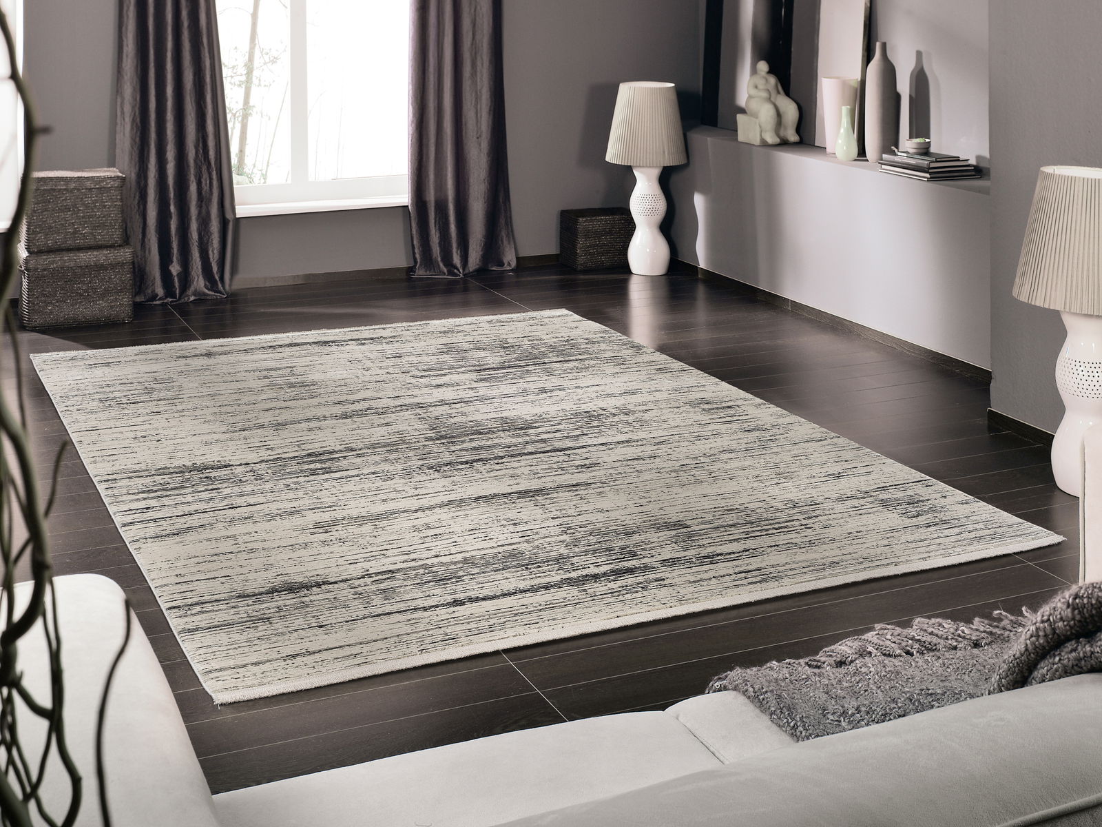 Moderner Designer Teppich in einem stilvollen Wohnzimmer, aus der Perspektive eines Sofas fotografiert. Der Teppich hat ein dezentes, graues Muster auf einem hellen Hintergrund und liegt auf einem dunklen Holzboden.