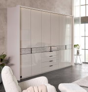 Grote, moderne kledingkast in hoogglans wit met meerdere deuren, een ladeblok in het midden en een decoratieve dwarsstreep, gefotografeerd vanuit een licht schuin perspectief van voren in een lichte slaapkamer.