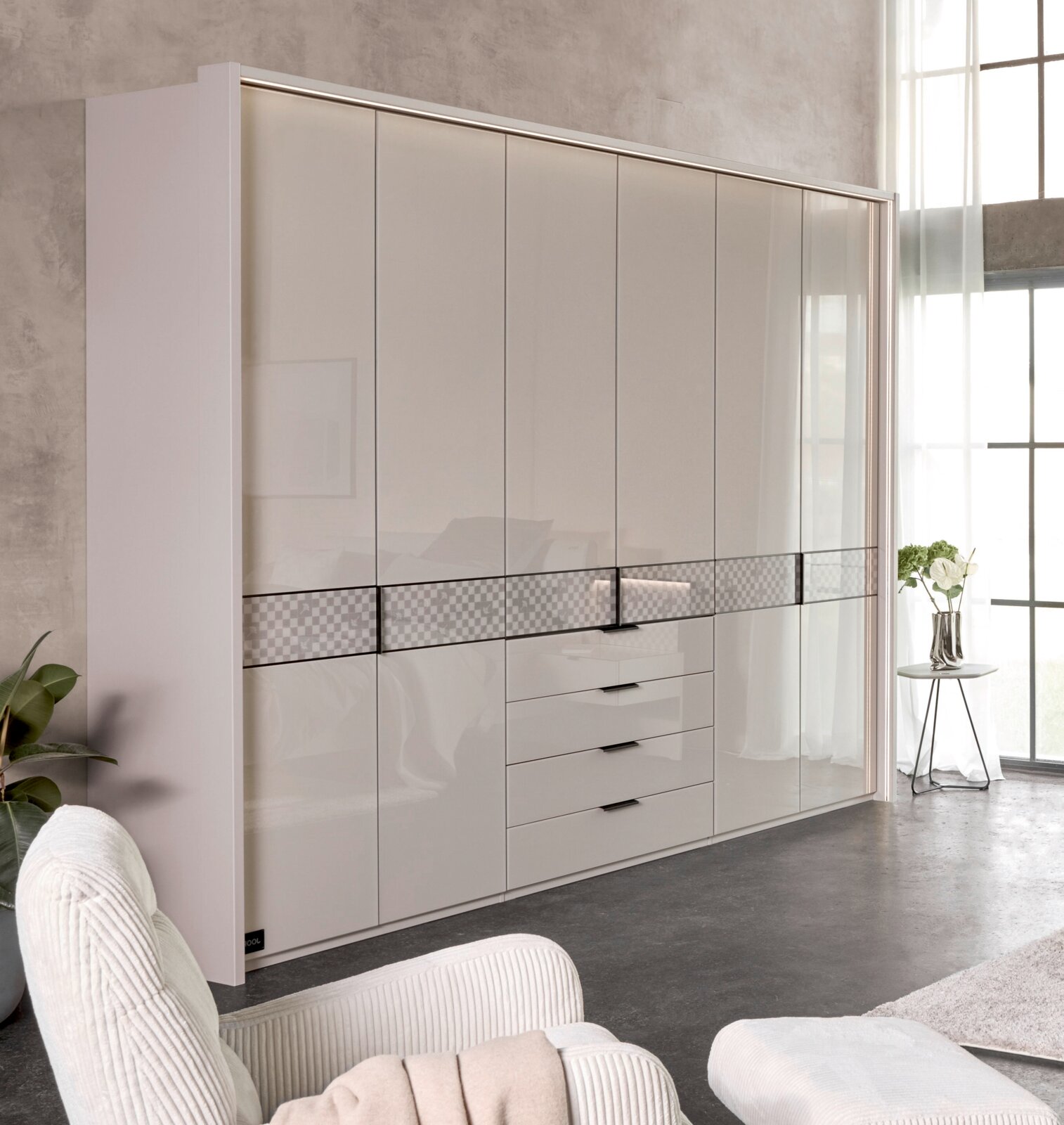 Grote, moderne kledingkast in hoogglans wit met meerdere deuren, een ladeblok in het midden en een decoratieve dwarsstreep, gefotografeerd vanuit een licht schuin perspectief van voren in een lichte slaapkamer.