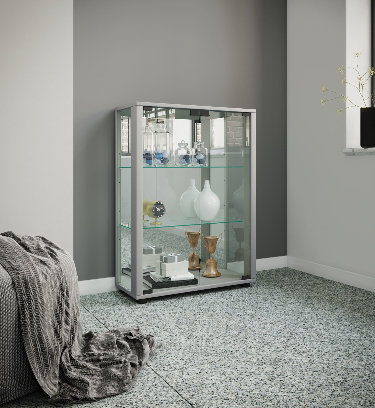 Moderne Glasvitrine mit silberfarbenem Rahmen und Glasschiebetüren, bestückt mit Deko-Objekten, in einem Wohnraum vor grauer Wand, Perspektive: frontale leicht seitliche Ansicht