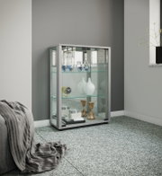 Moderne Glasvitrine mit silberfarbenem Rahmen und Glasschiebetüren, bestückt mit Deko-Objekten, in einem Wohnraum vor grauer Wand, Perspektive: frontale leicht seitliche Ansicht