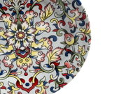 Draufsicht auf einen runden Porzellanteller mit farbenfrohem, ornamentalem Blumen- und Rankenmuster in Rot, Blau, Gelb und Grün auf hellem Hintergrund.