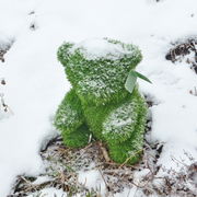 Grüner Bär aus künstlichem Gras, frontal im Schnee sitzend, mit einer leichten Schneeschicht bedeckt.