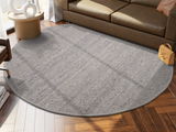 Runder, grauer Handwebteppich in einem modernen Wohnzimmer, auf einem Holzboden vor einem braunen Sofa platziert. Perspektive von oben.