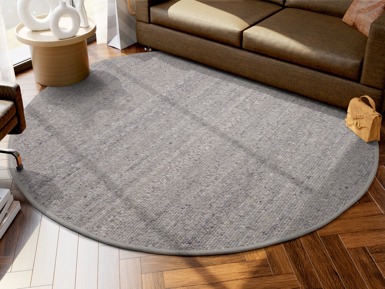 Runder, grauer Handwebteppich in einem modernen Wohnzimmer, auf einem Holzboden vor einem braunen Sofa platziert. Perspektive von oben.