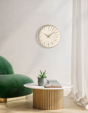 Moderne Wanduhr mit goldenen Akzenten an einer weißen Wand, im Hintergrund ein grüner Sessel und ein runder Tisch mit Pflanze, aus seitlicher Perspektive.