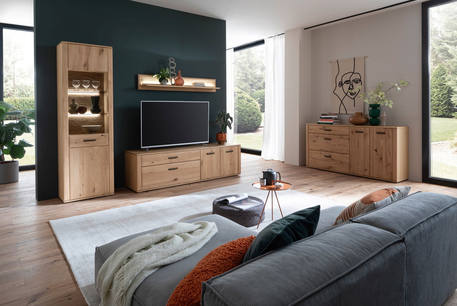 Moderne Wohnzimmermöbelkombination aus Holz, bestehend aus einem hohen Schrank mit Glaseinsätzen, einem TV-Board und einem Sideboard, in einem hellen Raum mit großen Fenstern und grüner Aussicht. Perspektive von der Seite des Sofas.
