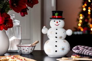 Porzellandose in Form eines Schneemanns mit schwarzem Zylinder, verziert mit bunten Schneeflocken, auf einem Tisch in weihnachtlicher Umgebung, frontal fotografiert.