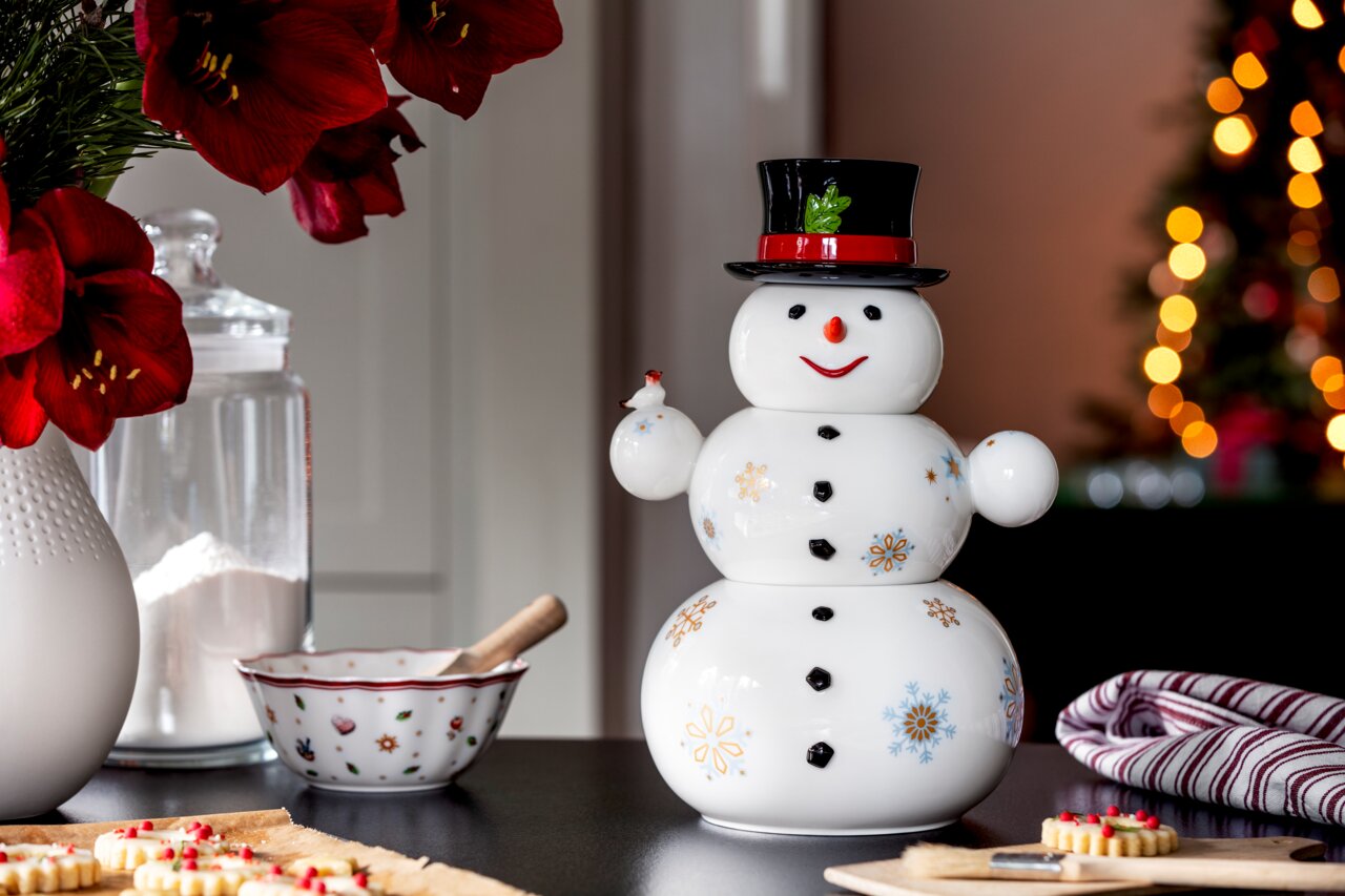 Porzellandose in Form eines Schneemanns mit schwarzem Zylinder, verziert mit bunten Schneeflocken, auf einem Tisch in weihnachtlicher Umgebung, frontal fotografiert.