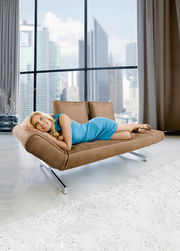 Frau in blauem Kleid liegt entspannt auf einem braunen Funktionssofa mit Metallfüßen, seitliche Perspektive