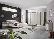 Modernes Schlafzimmer mit einem eleganten weißen Bett, seitlicher Perspektive. Das Bett ist mit Kissen und einer roten Decke dekoriert, umgeben von stilvollen Möbeln und Dekorationen.