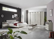 Modernes Schlafzimmer mit einem eleganten weißen Bett, seitlicher Perspektive. Das Bett ist mit Kissen und einer roten Decke dekoriert, umgeben von stilvollen Möbeln und Dekorationen.