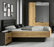 Modernes Schlafzimmer mit einem Bett, das einen Sockelschubkasten integriert hat. Das Bett ist aus Holz mit dunklen Akzenten und steht in einem stilvoll eingerichteten Raum mit grauen Wänden und dekorativen Bildern. Perspektive von der Seite.