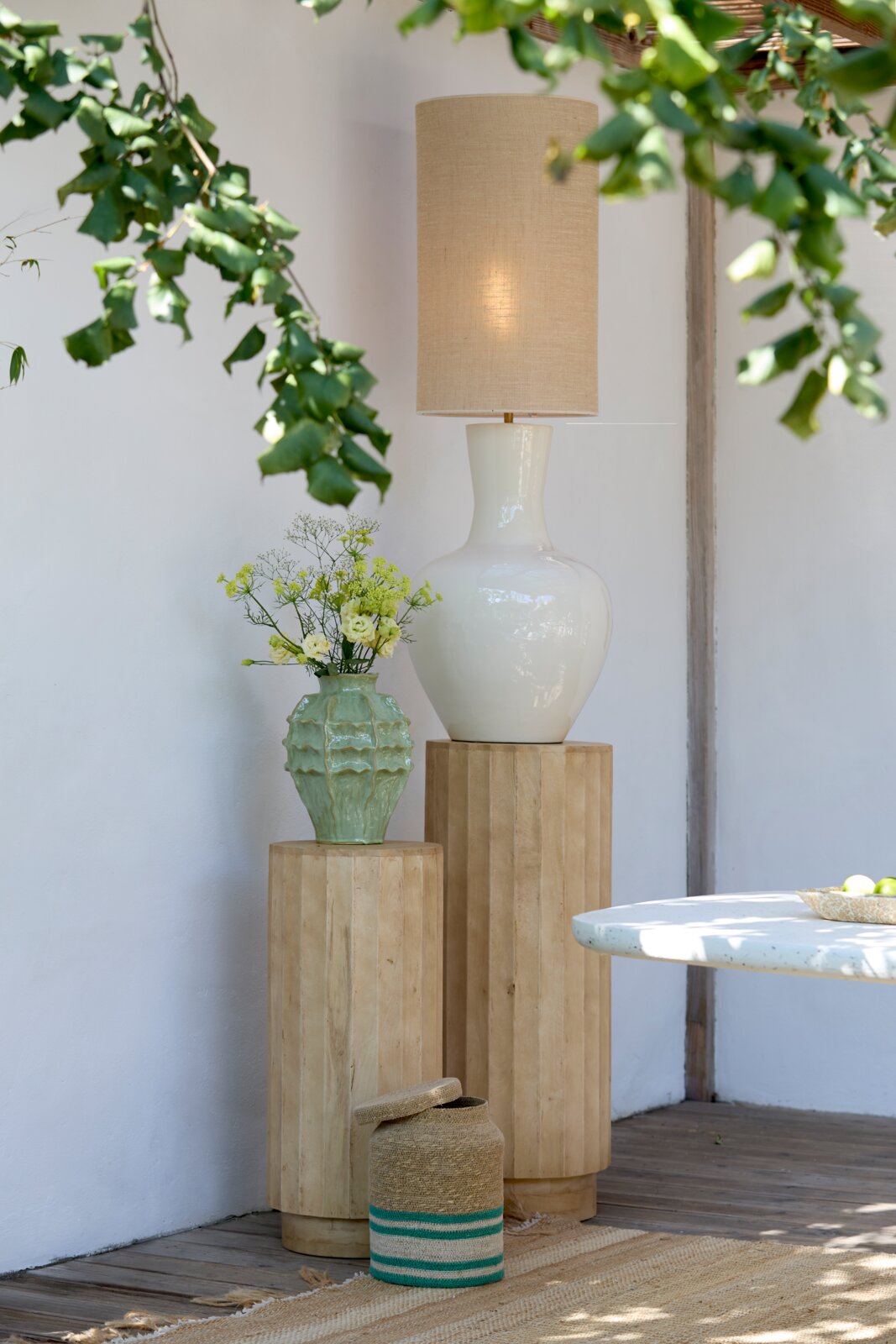 Frontale Innenraumaufnahme: weiße Keramik-Tischlampe mit beigem Stoffschirm auf hohem Holzpodest, daneben grüne Vase mit Blumen auf niedrigerem Podest Blätter im Vordergrund.