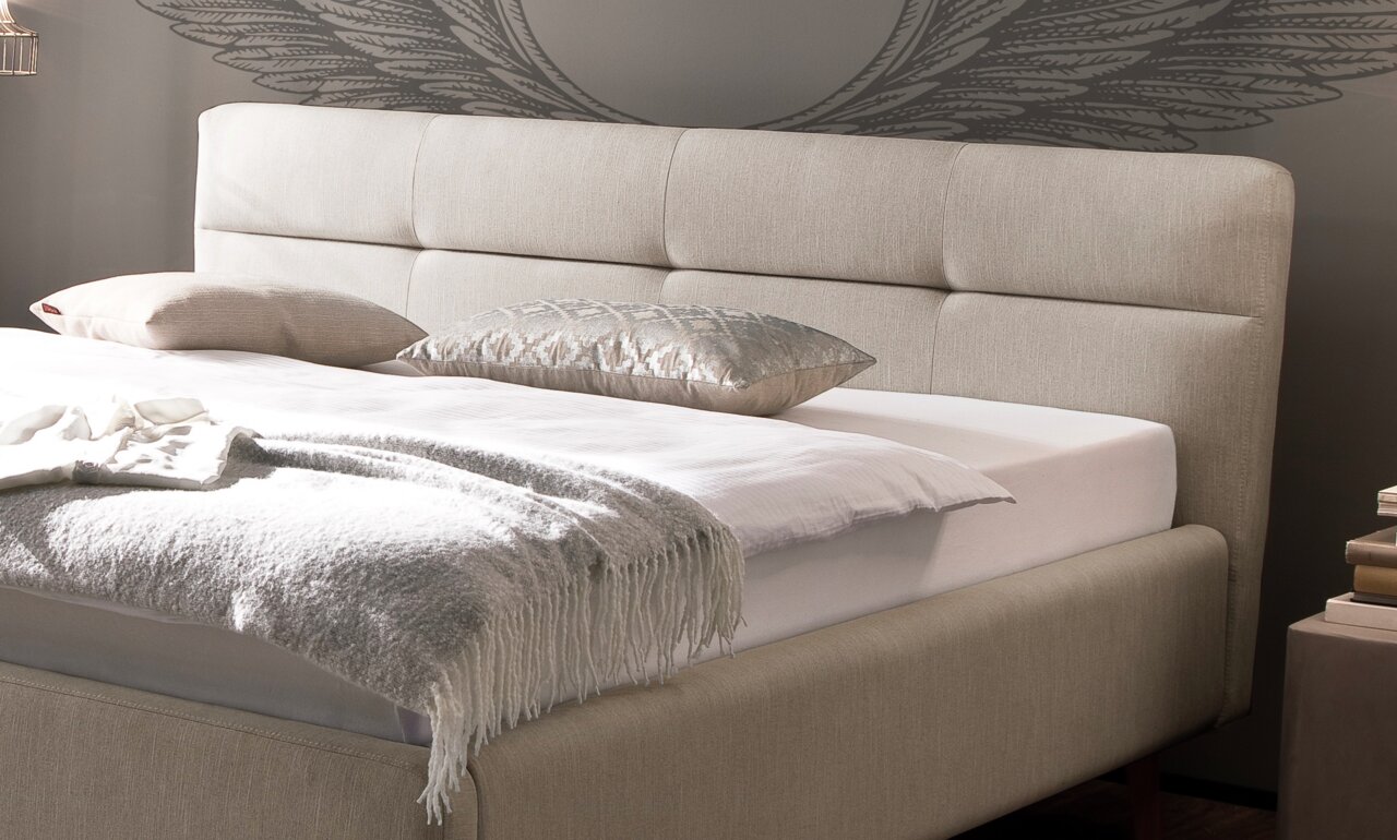 Beiges Polsterbett mit Kopfteil, seitlicher Blickwinkel, dekoriert mit Kissen und Decke.