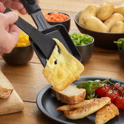 Nahaufnahme eines Raclette-Grills mit geschmolzenem Käse, der auf eine Brotscheibe gegossen wird. Im Hintergrund sind Schalen mit Gemüse und Kartoffeln zu sehen.