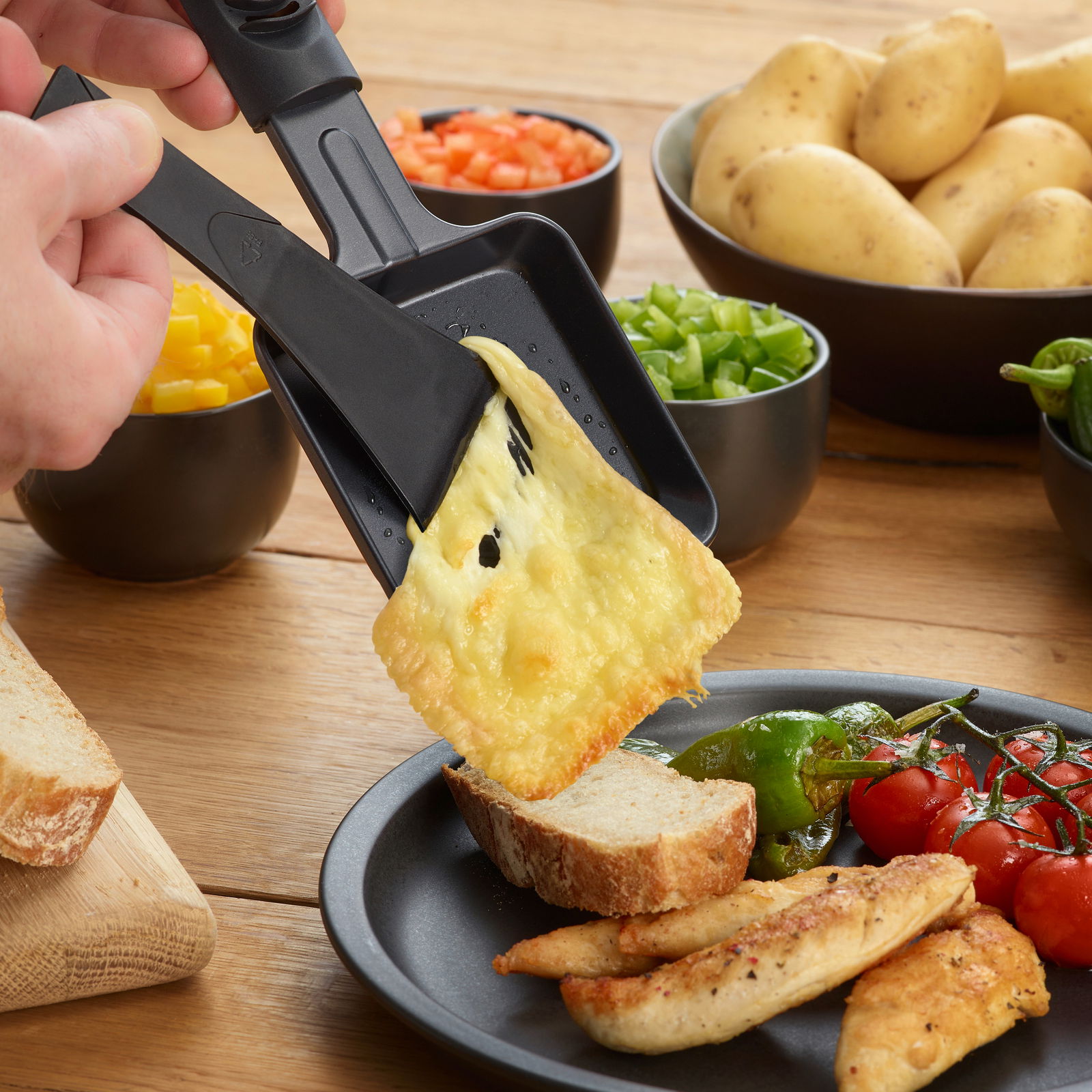 Nahaufnahme eines Raclette-Grills mit geschmolzenem Käse, der auf eine Brotscheibe gegossen wird. Im Hintergrund sind Schalen mit Gemüse und Kartoffeln zu sehen.