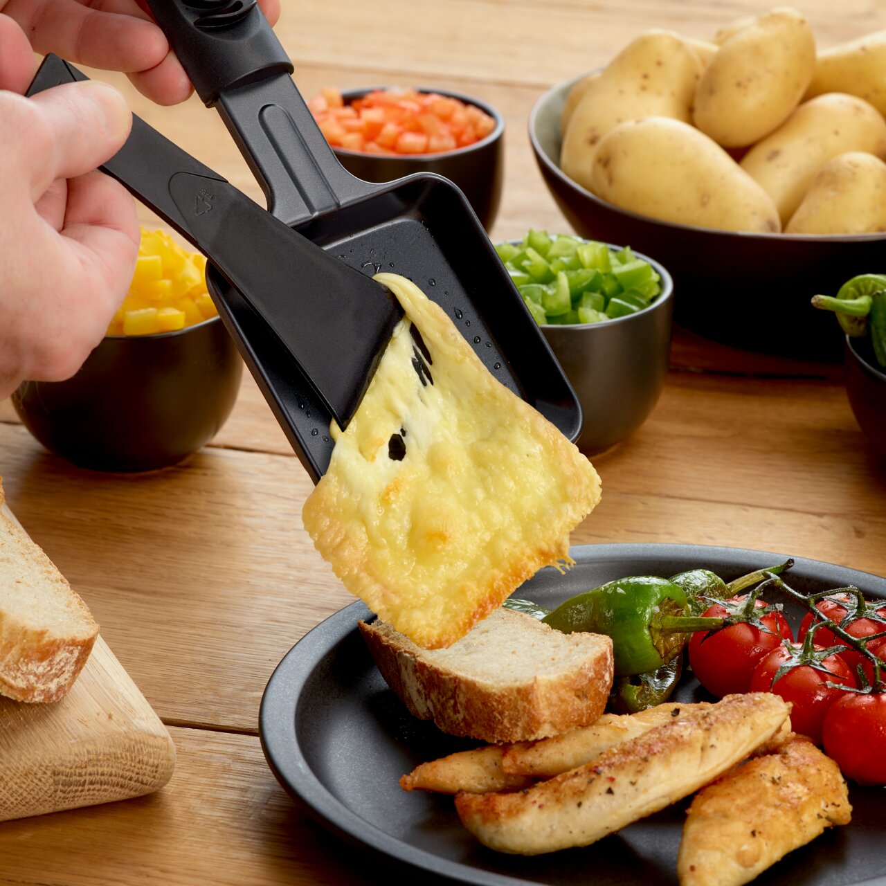 Nahaufnahme eines Raclette-Grills mit geschmolzenem Käse, der auf eine Brotscheibe gegossen wird. Im Hintergrund sind Schalen mit Gemüse und Kartoffeln zu sehen.