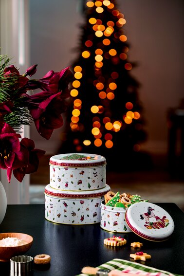Drei festliche Keksdosen mit weihnachtlichen Motiven, gestapelt auf einem Tisch vor einem unscharfen Weihnachtsbaum im Hintergrund, aus einer seitlichen Perspektive fotografiert.