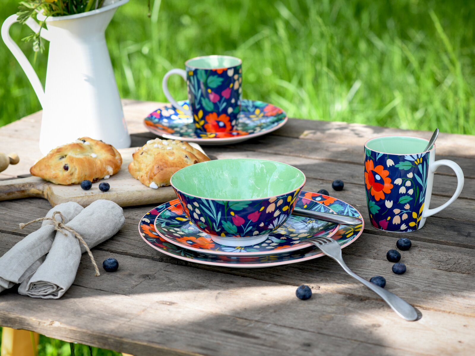Buntes Geschirrset mit floral gemusterten Tassen, Schale und Tellern auf rustikalem Holztisch im Garten, mit Blaubeeren und Gebäck, fotografiert in leichter Draufsicht/Schrägansicht