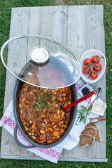 Alugussbräter Alucast 38cm mit Glasdeckel, gefüllt mit einem Eintopf aus Bohnen und Gemüse, von oben fotografiert. Daneben liegen Tomaten, Pfeffer, Gewürze und Brotscheiben auf einem Holztisch im Freien.