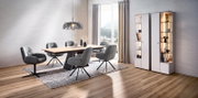 Modernes Esszimmer-Set mit rechteckigem Holztisch, vier grauen Polsterstühlen auf Teppich, Pendelleuchte und beleuchteten weißen Vitrinenschränken neben bodentiefem Fenster, Perspektive: leicht schräg von vorn aus dem Raum