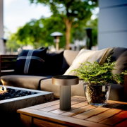 Moderne, zylindrische Outdoor-Tischlampe in Anthrazit auf einem Holztisch neben einer Pflanze, im Hintergrund eine Lounge im Garten; Perspektive: Nahaufnahme aus leicht erhöhter, frontaler Ansicht.