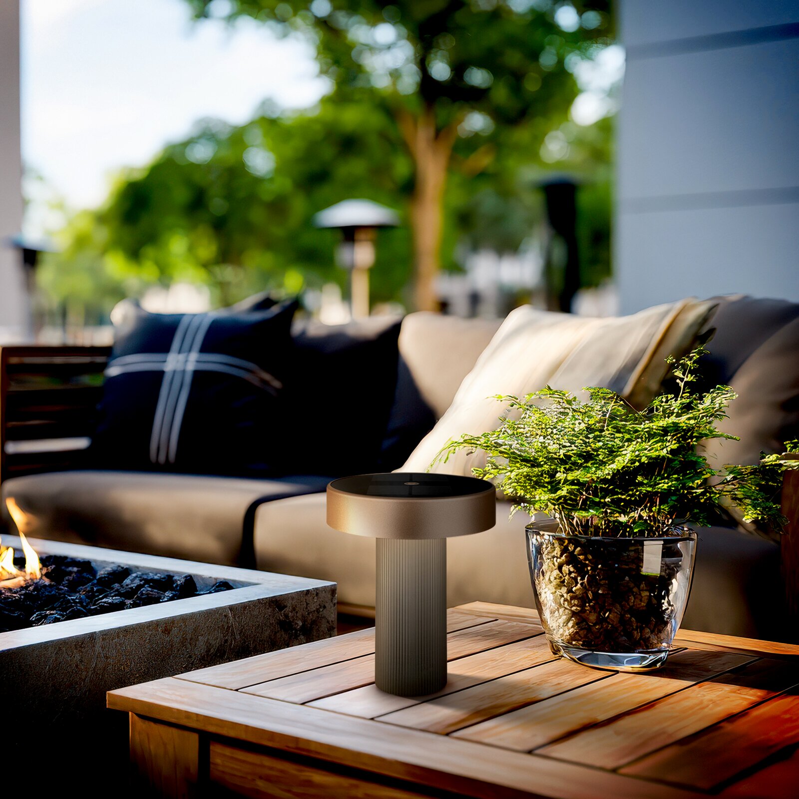 Moderne, zylindrische Outdoor-Tischlampe in Anthrazit auf einem Holztisch neben einer Pflanze, im Hintergrund eine Lounge im Garten Perspektive: Nahaufnahme aus leicht erhöhter, frontaler Ansicht.