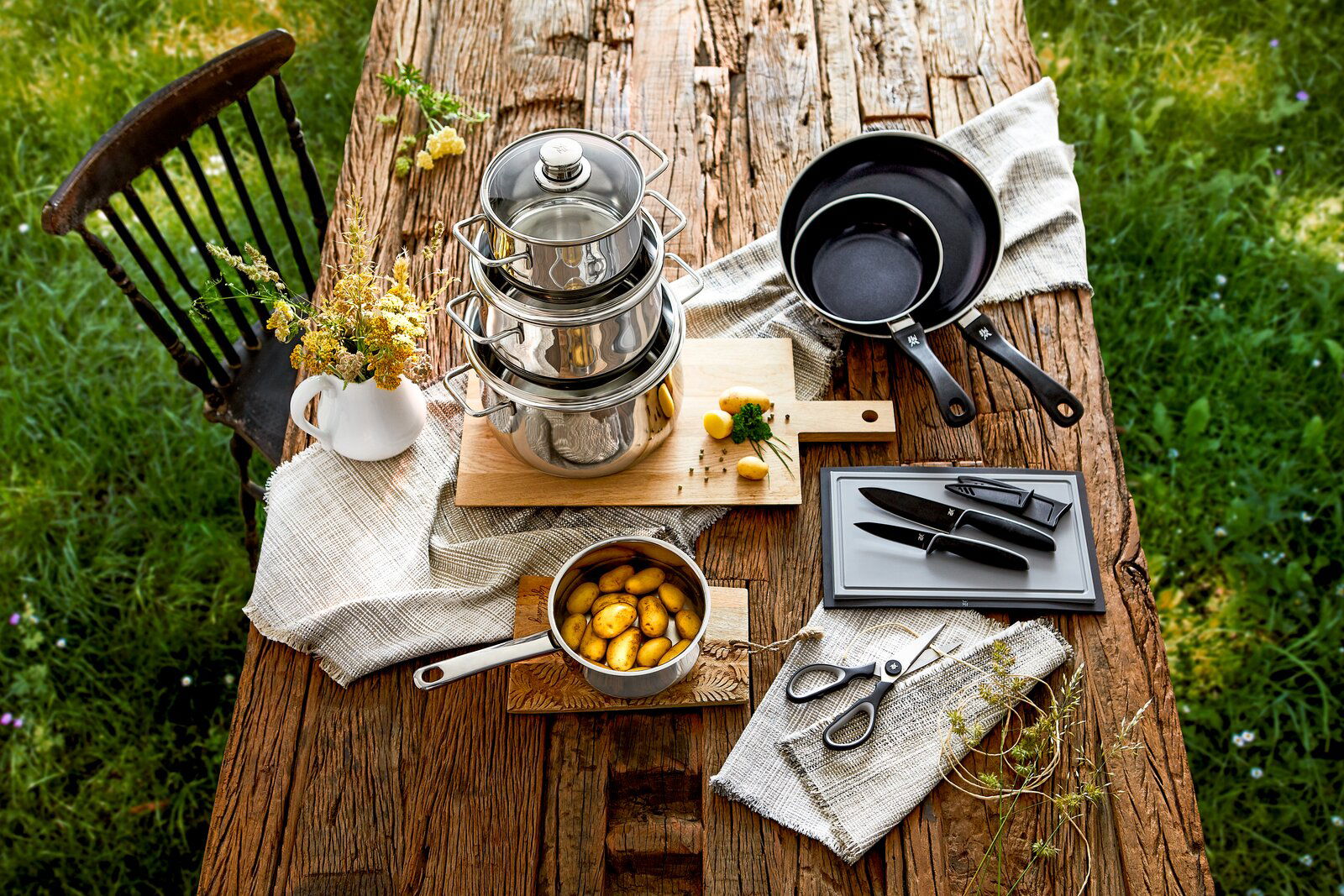 Kochtopf-Starter-Set 10-tlg. auf einem rustikalen Holztisch, bestehend aus gestapelten Edelstahltöpfen, Pfannen, einem Topf mit Kartoffeln, Messern und einer Schere, aus der Vogelperspektive fotografiert.