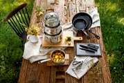 Kochtopf-Starter-Set 10-tlg. auf einem rustikalen Holztisch, bestehend aus gestapelten Edelstahltöpfen, Pfannen, einem Topf mit Kartoffeln, Messern und einer Schere, aus der Vogelperspektive fotografiert.