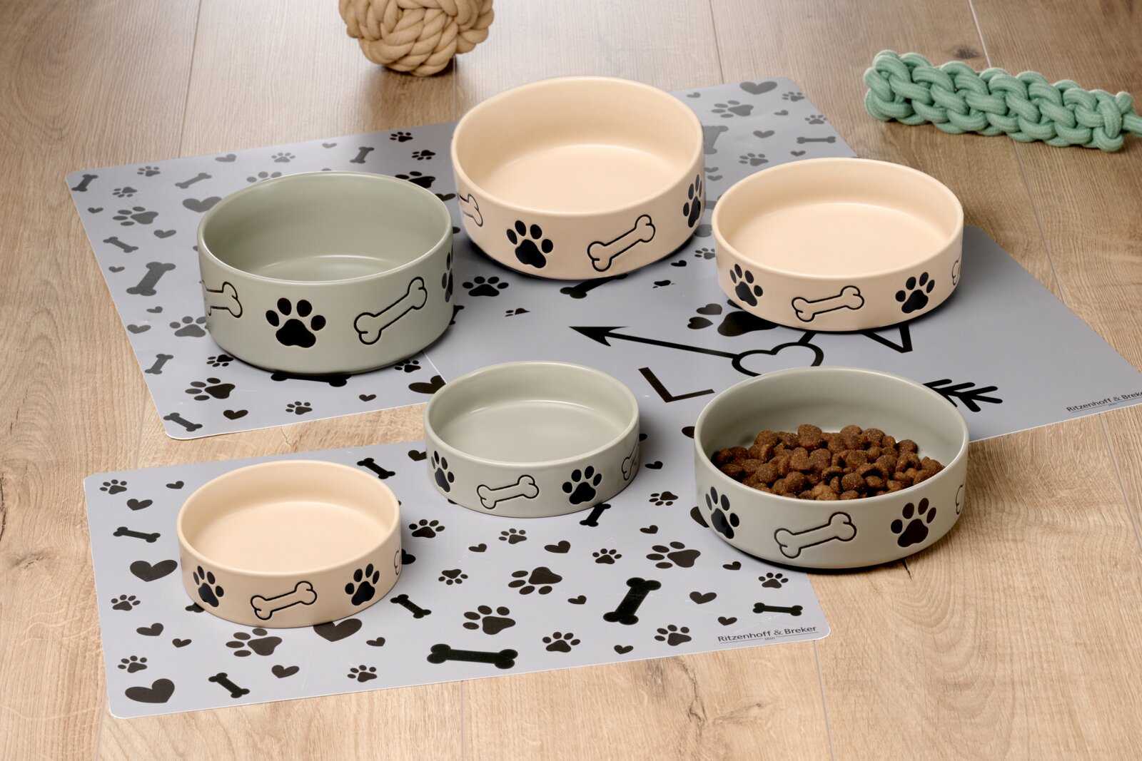 Verschillende hondenbakjes in beige en groen met pootafdrukken en bottenmotieven, gefotografeerd vanaf een schuine hoek bovenop een decoratieve ondergrond.