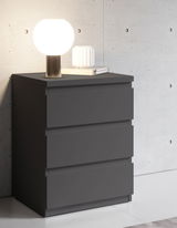 Moderne Nachtkommode in dunklem Grau mit drei Schubladen, darauf eine runde Tischlampe und ein Buch, seitliche Perspektive