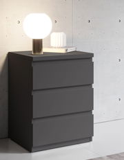 Moderne Nachtkommode in dunklem Grau mit drei Schubladen, darauf eine runde Tischlampe und ein Buch, seitliche Perspektive