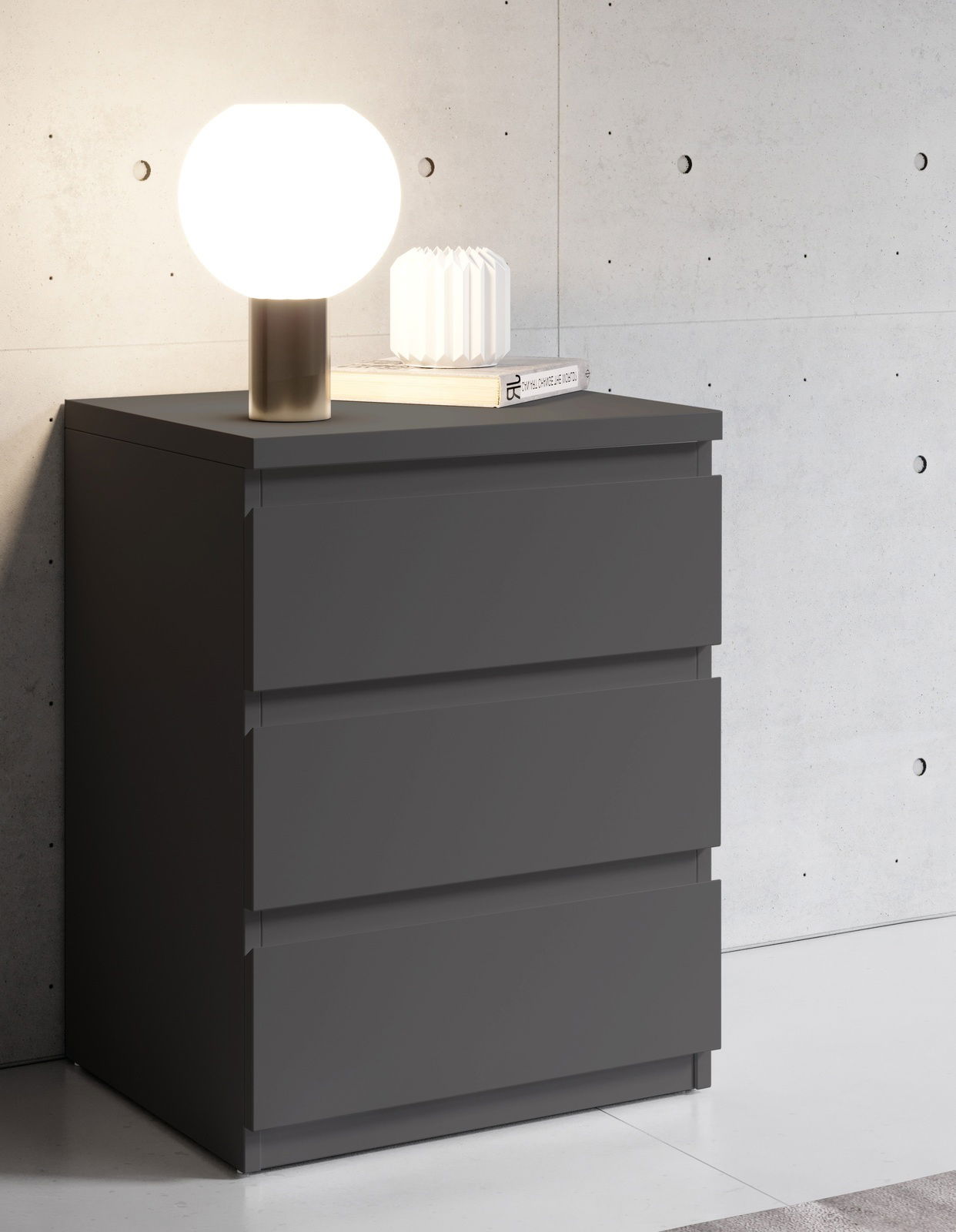 Moderne Nachtkommode in dunklem Grau mit drei Schubladen, darauf eine runde Tischlampe und ein Buch, seitliche Perspektive