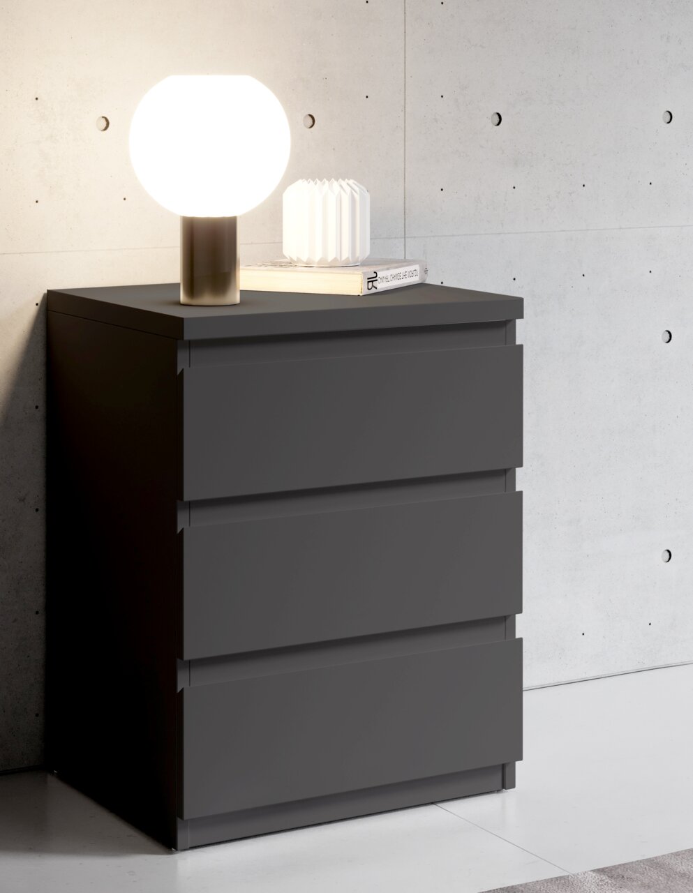 Moderne Nachtkommode in dunklem Grau mit drei Schubladen, darauf eine runde Tischlampe und ein Buch, seitliche Perspektive