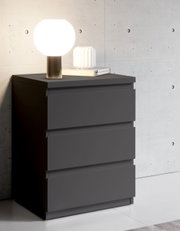 Moderne Nachtkommode in dunklem Grau mit drei Schubladen, darauf eine runde Tischlampe und ein Buch, seitliche Perspektive