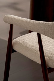 Nahaufnahme eines gepolsterten Stuhls mit cremefarbenem Bouclé-Bezug und dunkelbraunem Holzgestell, perspektivisch von der Seite leicht von oben fotografiert.