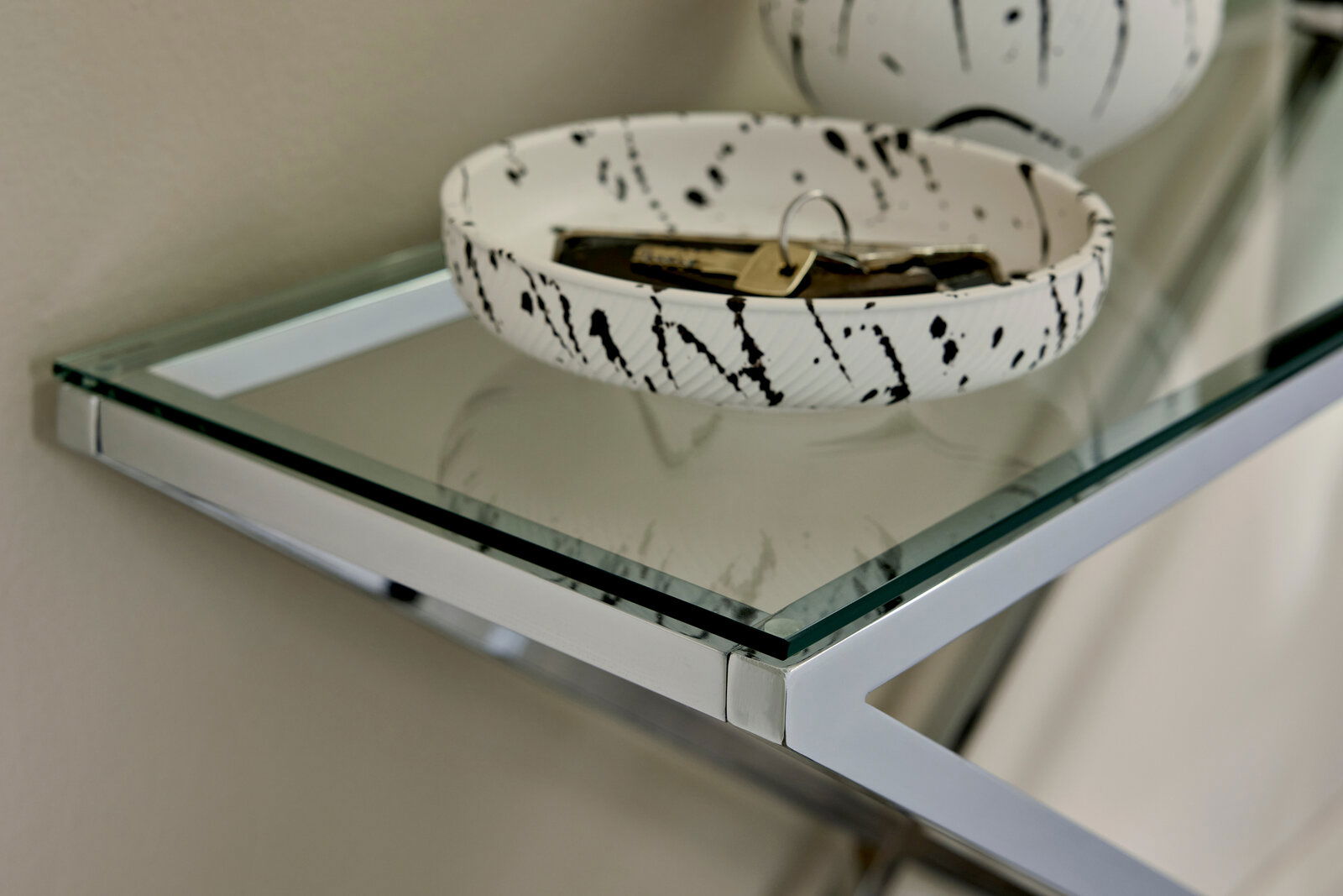 Nahaufnahme einer modernen Konsole mit Glasplatte und silbernem Metallrahmen, darauf eine dekorative Schale mit Schlüsseln, seitliche Perspektive.