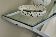 Nahaufnahme einer modernen Konsole mit Glasplatte und silbernem Metallrahmen, darauf eine dekorative Schale mit Schlüsseln, seitliche Perspektive.
