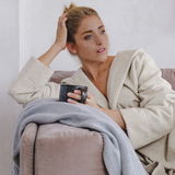 Frau in beigem Kapuzenbademantel sitzt entspannt auf einem Sofa und hält eine Tasse, seitliche Perspektive